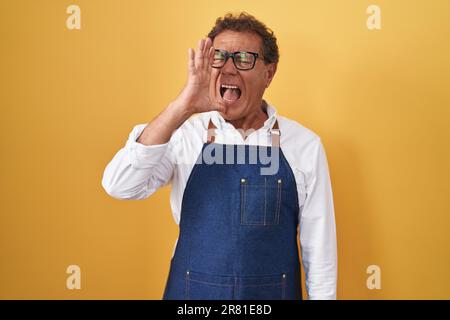 Ein hispanischer Mann mittleren Alters, der eine professionelle Kochschürze trägt, schreit und schreit laut zur Seite mit der Hand auf dem Mund. Kommunikationskonzept. Stockfoto