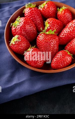 Frische reife Erdbeere in Schüssel auf dunklem Hintergrund. Tonschüssel mit Erdbeeren auf blauer Serviette. Speicherplatz kopieren. Draufsicht Stockfoto