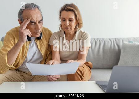 Haushaltspapiere. Seriöse pensionierte Ehepartner, die ihren Laptop benutzen, Rechnungen lesen, Dokumente durchsehen, auf der Couch sitzen Stockfoto