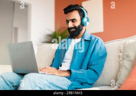 Glücklicher inder, der zu Hause Computer und kabellose Kopfhörer benutzt Stockfoto