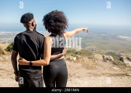 Eine afroamerikanische Dame umarmt einen Mann, genießt Trainingspause, zeigt den Finger zur Seite, steht auf Felsen am Meer, draußen Stockfoto