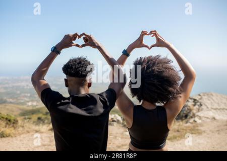 Ein junges, schwarzes, sportliches Paar, das draußen trainiert und mit den Händen ein Schild mit Herz macht, auf Felsen am Meer steht, Blick nach hinten Stockfoto