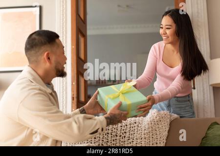Eine Asiatin, die einem Mann im Wohnzimmer eine Geschenkbox schenkt Stockfoto