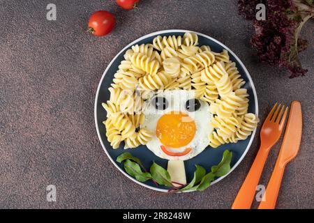 Lustiges Essen für Kinder - lächelndes Gesicht von Spiegeleiern, Nudeln, Oliven und Salat auf blauem Teller. Kreatives, gesundes Frühstück für Kinder Stockfoto