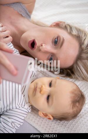 Eine Frau mit einem Baby, die ein Selfie macht, liegt auf dem Bett Stockfoto