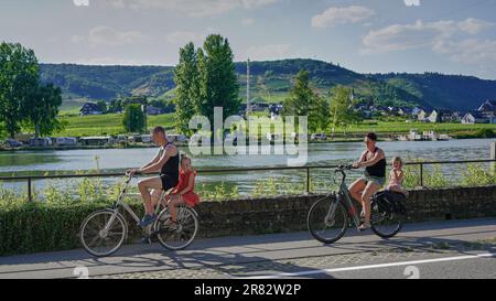 Familienausflug mit dem Fahrrad auf der Mosel in Deutschland. Mann, Frau und zwei junge Mädchen, die zwei Fahrräder auf dem Radweg neben dem ruhigen Wasser der Mosel fahren Stockfoto