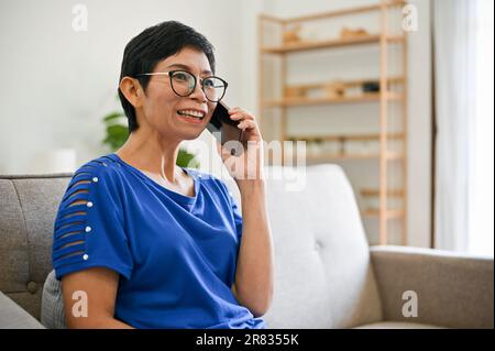 Eine fröhliche, reife Asiatin mittleren Alters, die mit ihrem Enkel telefoniert und sich auf dem Sofa in ihrem Wohnzimmer entspannt. Stockfoto