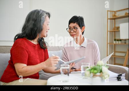 Eine glückliche und professionelle asiatische Krankenversichererin mittleren Alters besuchte eine Rentnerin zu Hause, um einen Versicherungsvertrag abzuschließen. Stockfoto