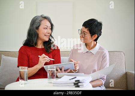 Eine schöne, reife asiatische Krankenversichererin mittleren Alters besuchte eine Rentnerin zu Hause, um Geschäfte zu machen und einen neuen Versicherungsvertrag abzuschließen. Stockfoto