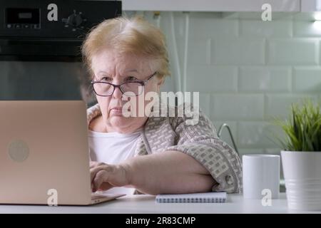 Fokussierte ältere Frau, die zu Hause am Tisch sitzt und mit einem Computer-Laptop mit ernstem Gesicht arbeitet Stockfoto
