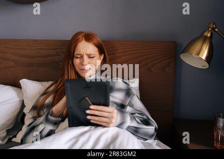 Porträt einer depressiven jungen Frau, die auf dem Bett liegt, ein Foto im Rahmen hält und weint, das Bild ansieht, das Foto mit Liebe berührt. Stockfoto