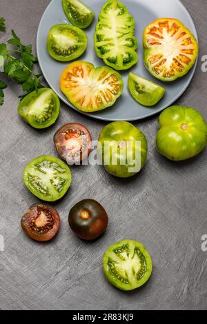 Verschiedene Sorten grüner und roter Tomaten auf einem Teller und auf dem Tisch. Flach verlegt. Grauer Hintergrund. Stockfoto