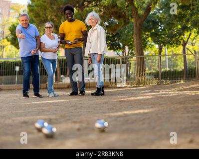 Metall-Petanque-Kugeln gegen vier verschwommene mehrrassische ältere Spieler in der Ferne im Freien Stockfoto