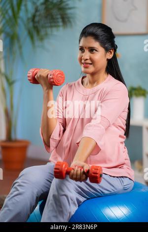 Vertikale Aufnahme einer glücklichen indischen schwangeren Frau, die Sport macht, indem sie Kurzhanteln benutzt, während sie zu Hause auf dem Fitnessball sitzt - Konzept eines gesunden Lebensstils Stockfoto