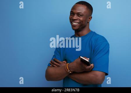30-jähriger, gutaussehender, mit dunkler Haut ausgestatteter amerikanischer Mann in einem einfachen T-Shirt hält ein Smartphone mit der Hand und lächelt Stockfoto