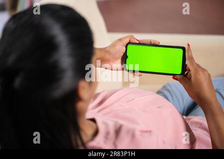 Schulterfoto einer indischen schwangeren Frau, die in ein Mobiltelefon mit grünem Bildschirm schaut, während sie zu Hause auf dem Sofa sitzt - Konzept von App-Werbung, Technologie und Stockfoto