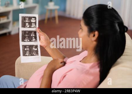 Schulterfoto einer glücklichen indischen schwangeren Frau auf dem Sofa, die auf einen Ultraschallbericht schaut, indem sie den Bauch zu Hause hält - Konzept der Schwangerschaftsreise, mütterlich Stockfoto