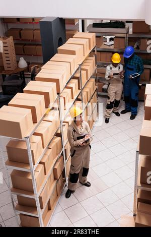 Lagerassistenten stehen in der Nähe von Kartonregalen und führen Inventuren durch. Kaukasische und afroamerikanische Kollegen tragen Schutzhelme, die im Lager für Lieferdienste arbeiten Stockfoto