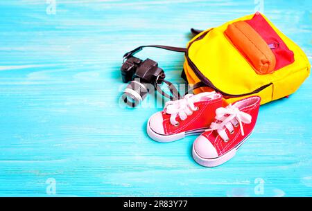 Set aus kleinen roten, hohen Segelschuhen, einem winzigen Rucksack und einer Spielzeugkamera auf blauem Holzhintergrund. Kreatives Back-to-School-Konzept mit t Stockfoto