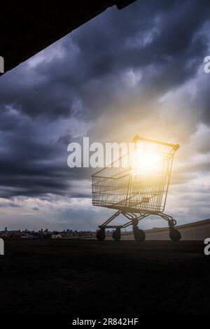 Ein Einkaufswagen auf einem Parkplatz auf dem Dach eines verlassenen Einkaufszentrums, der Sonnenuntergang durch Regenwolken im Hintergrund. Stockfoto