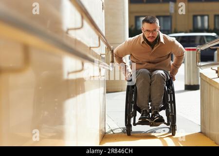 Junger Mann im Rollstuhl, der die Rampe des Gebäudes hochfährt Stockfoto