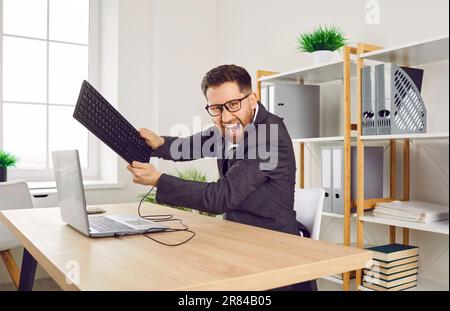 Verärgerter Geschäftsmann am Arbeitsplatz im Büro, der versucht, den Monitor mit seiner Tastatur zu zerstören. Stockfoto