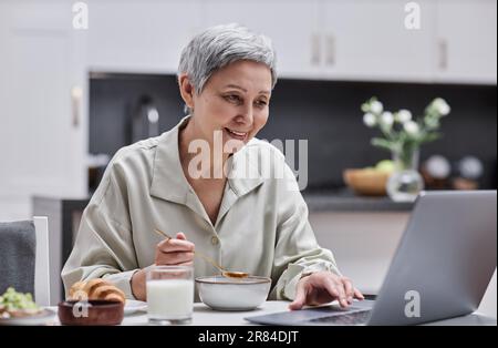 Porträt einer lächelnden Seniorin, die ihren Laptop benutzt und arbeitet, während sie zu Hause frühstückt, im Kopierbereich Stockfoto