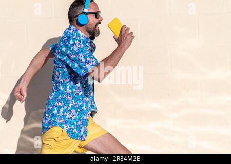 Junger bärtiger Hipster in einem hawaiianischen Hemd, einer Sonnenbrille und kabellosen Kopfhörern läuft, während er lächelt. Hält sein gelbes Smartphone in der Hand.copy s Stockfoto