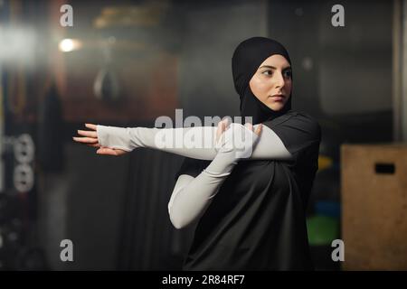 Junge muslimische Sportlerin in Hijab und Sportbekleidung streckt den linken Arm, während sie beim Fitnesstraining im Fitnessstudio vor der Kamera steht Stockfoto