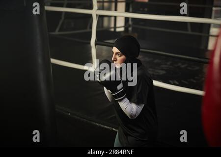 Junge, ernsthafte muslimische Boxerin in Aktivbekleidung und Hijab, die beim Training auf einen Boxsack schaut und sich auf den weiteren Schlag konzentriert Stockfoto