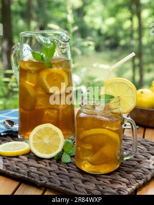 Eistee im Glasgefäß und Krug mit Zitrone und Minze im Freien auf einem Holztisch mit Naturhintergrund Stockfoto