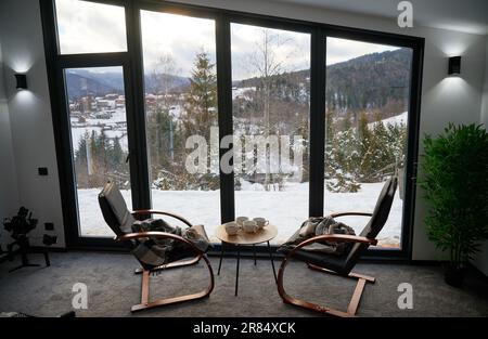Gemütliches Zimmer mit kleinem Couchtisch und Sesseln vor einem großen Panoramafenster. Vorderansicht von zwei Holzstühlen und Tisch vor einem großen Fenster mit verschneiter Berglandschaft. Urlaubskonzept. Stockfoto