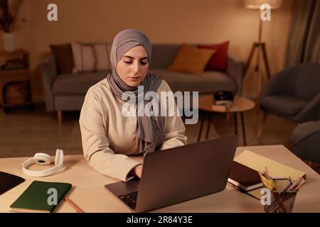 Junge, ernsthafte muslimische Geschäftsfrau, die am Arbeitsplatz im Wohnzimmer sitzt und auf den Laptop-Bildschirm schaut, während sie durch Online-Daten scrollt Stockfoto