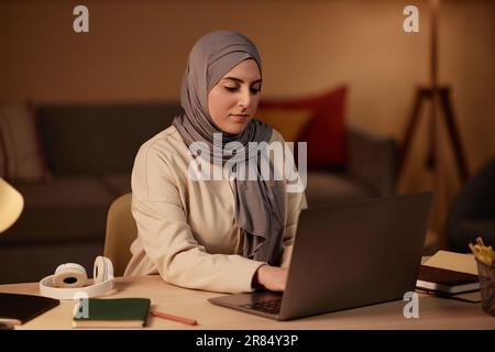 Junge muslimische Geschäftsfrau in Hidschab- und Freizeitkleidung, die auf einer Laptop-Tastatur tippt, während sie am Arbeitsplatz im Home Office sitzt und Netzwerke nutzt Stockfoto