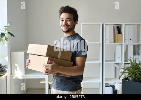 Porträt eines jungen Mannes, der vor der Kamera lächelt, während er im Büro verpackte Kartons transportiert Stockfoto