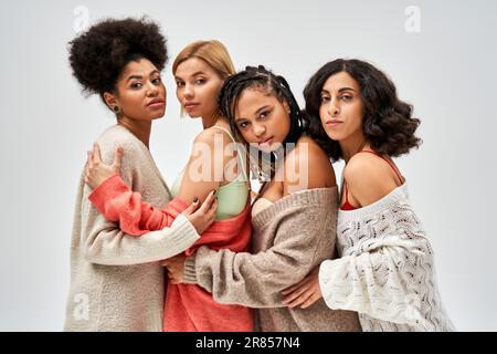 Porträt von selbstbewussten und multiethnischen Frauen in Strickpullover, die sich umarmen und in die Kamera schauen, isoliert auf grau, verschiedenen Körpertypen und selbstsüchtig Stockfoto