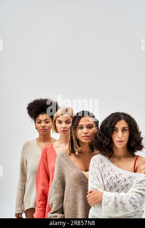 Multiethnische Frauen in warmen Strickpullis, die in die Kamera schauen und nebeneinander stehen, isoliert auf Grau, verschiedene Körpertypen und selbstgefällig Stockfoto