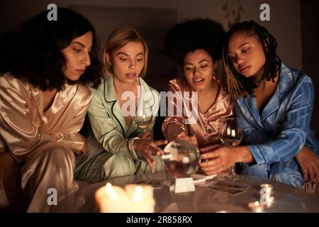 Multiethnische Frauen in farbenfrohem Pyjama, die Gläser Wein halten und in der Nacht der Mädchen zu Hause eine Kristallkugel in der Nähe von Tarotkarten und Kerzen anschauen Stockfoto