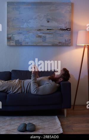Mobile Interaktion, bärtiger Mann mit roten Haaren mit Smartphone, ruht sich auf dem Sofa, malt an der Wand, Hausschuhe auf Teppich, Nacht, Licht aus der Lampe, Freizeit Stockfoto