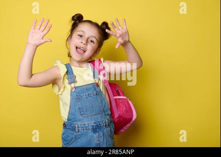 Fröhliches Vorschulmädchen mit Rucksack, zeigt vor der Kamera ihre Handflächen, lächelt und genießt den Beginn des neuen Schuljahres Stockfoto