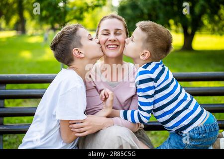 Glückliche Mutter sitzt mit ihren Söhnen auf einer Bank im Park. Sie haben Spaß zusammen. Die Jungs küssen ihre Mutter. Stockfoto