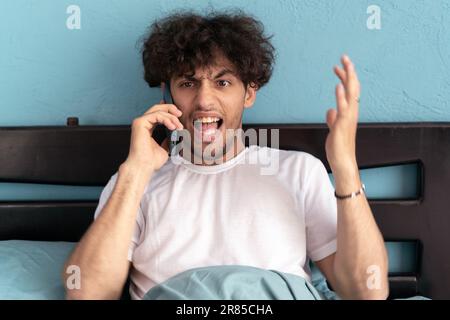 Junger araber, der im Bett sitzt, telefoniert und laut ins Telefon schreit, nervöser Stress Stockfoto