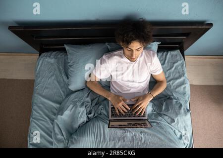 Junger arabischer Mann, der im Bett liegt und online mit einem Laptop arbeitet, online lernt, Podcast auf einem Laptop ansieht, Lernkurs anhört, Konferenz Stockfoto