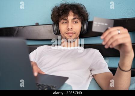 Junger, arabischer, glücklicher Mann im lässigen Pyjama-Outfit, der sitzt und die Kreditkarte auf dem Bett im Schlafzimmer hält, surft im Internet und kauft über den Laptop ein Stockfoto