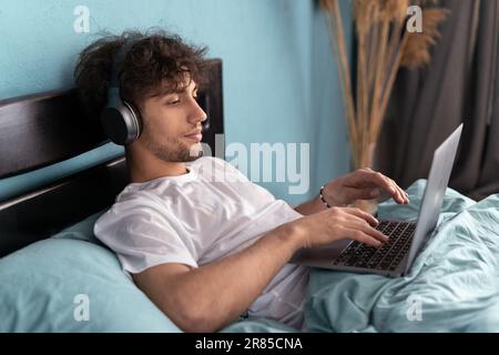 Ein Mann streamt einen Film in seinem Schlafzimmer über das Internet, während er entspannt ist. Glücklicher Mann mit Kopfhörern, der sich morgens zu Hause auf dem Computer-pc Serien ansieht Stockfoto