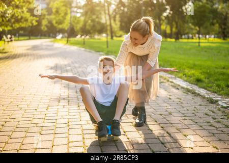Mutter hat Spaß mit ihrem Sohn im Park an sonnigen Tagen. Der Junge reitet Skateboard und seine Mutter schubst ihn. Stockfoto