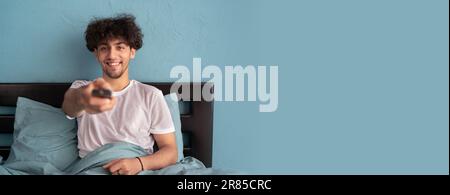 Ein junger arabischer Mann, der auf dem Bett sitzt, während er den Fernsehsender umschaltet, benutzt die Fernbedienung. Stockfoto