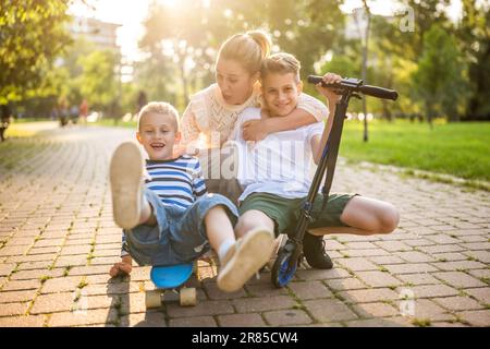Fröhliche Mutter, die Spaß mit ihren beiden Söhnen im Park an sonnigen Tagen hat. Sie reiten Skateboard und Roller. Stockfoto
