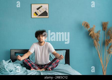 Junger Mann, der Yoga im Bett praktiziert, morgens aufwacht. Meditationskonzept. Stockfoto