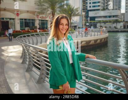 Junge Frau im grünen Anzug geht durch die Straßen von Dubai, Dubai Marina District. Reisekonzept der Vereinigten Arabischen Emirate. Die Vorstellung von erfolgreichem Auswanderer, Umzug in ein anderes Land, Arbeitsvisum Stockfoto
